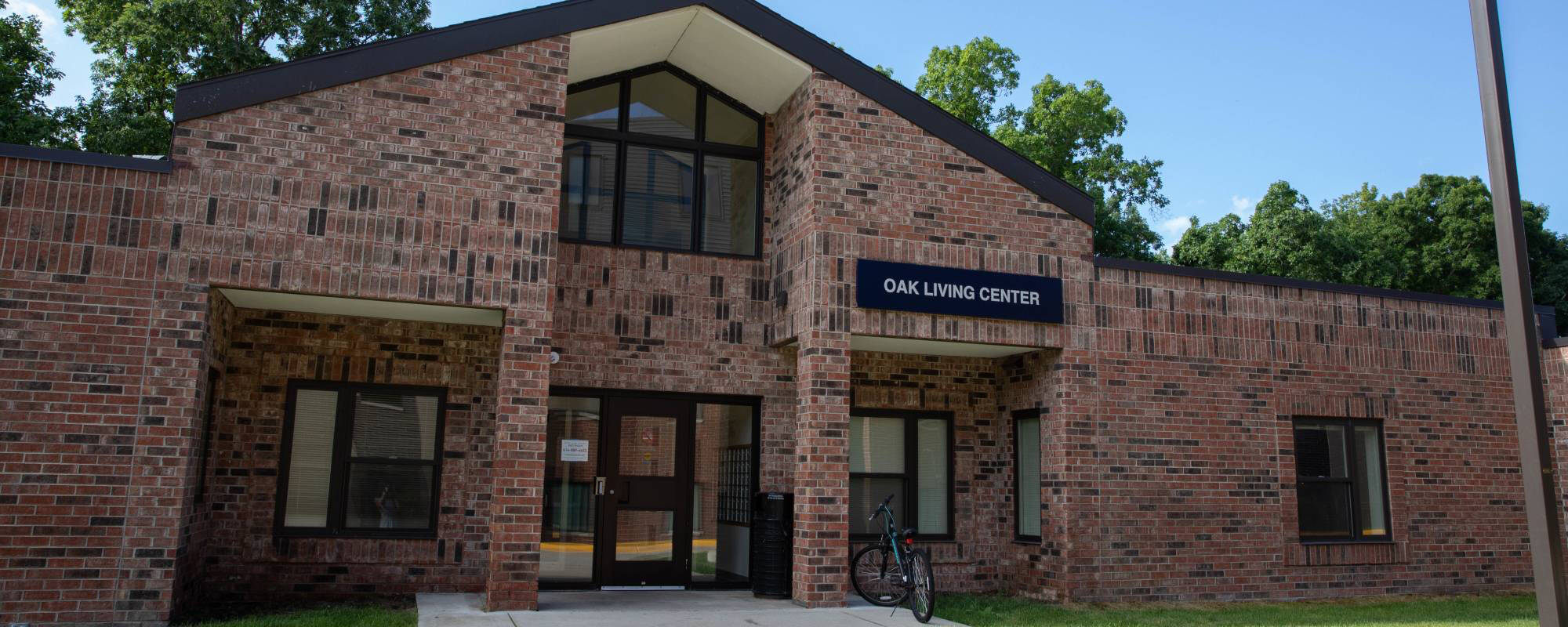 A brick building labeled "Oak Living Center" with a brown roof and large windows. A bicycle is parked at the entrance. The scene is calm and sunny.